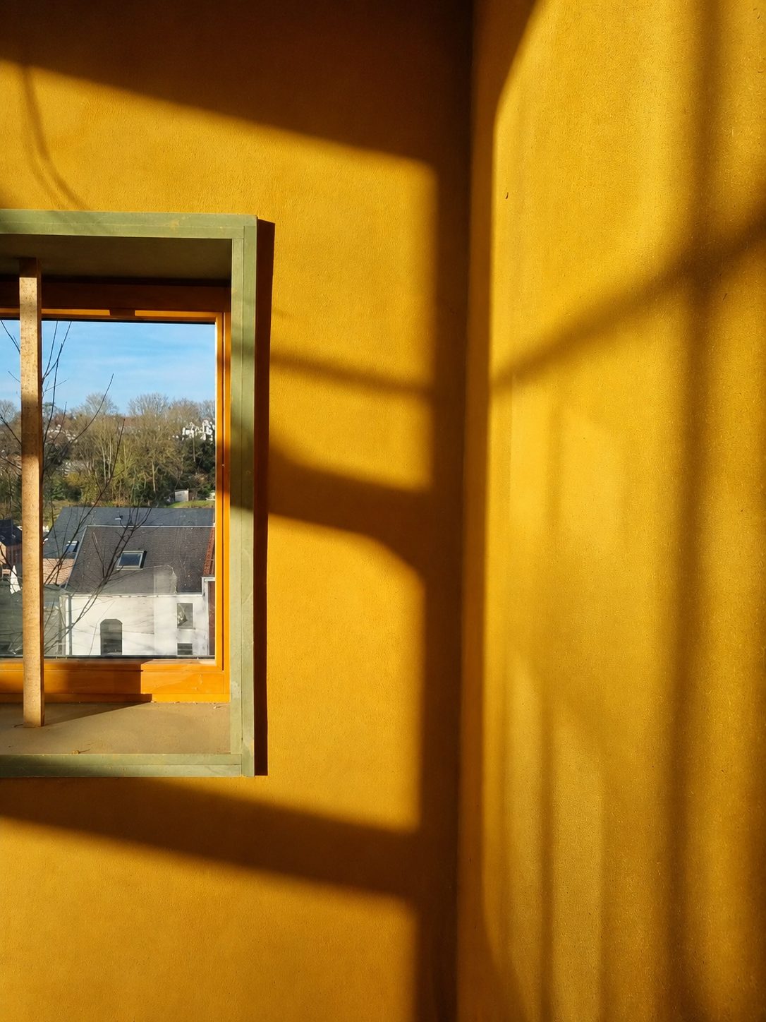 Mur en enduit terre ocre, lumière du matin et fenêtre à cadre vert